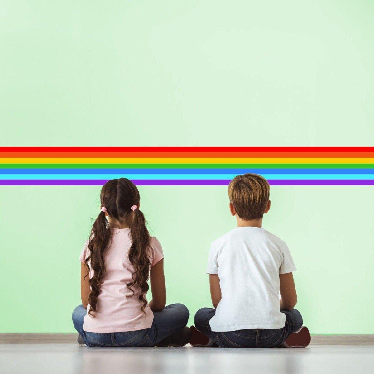 Two children sitting on the floor, admiring a vibrant rainbow stripe wall decal on light green wall.